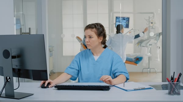 Dentist works at computer while dentist is in exam room behind her 457951541