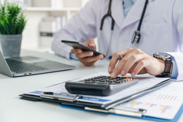  doctor using a calculator with healthcare metrics on a clipboard 659759183-1